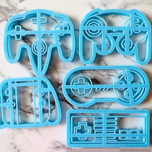 May include: Set of four blue plastic cookie cutters shaped like video game controllers. The cutters are in the shape of a Nintendo 64 controller, a Super Nintendo controller, a Game Boy, and a NES controller.