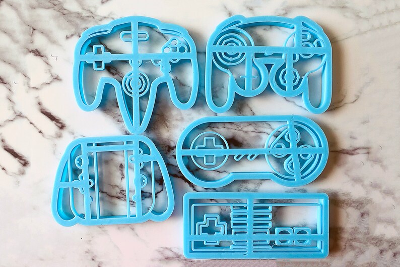 May include: Set of four blue plastic cookie cutters shaped like video game controllers. The cutters are in the shape of a Nintendo 64 controller, a Super Nintendo controller, a Game Boy, and a NES controller.