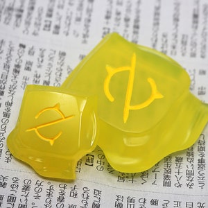May include: Two translucent yellow decorative pieces with a yellow symbol. The pieces are on a newspaper background. The symbol appears to be a stylized letter or emblem.