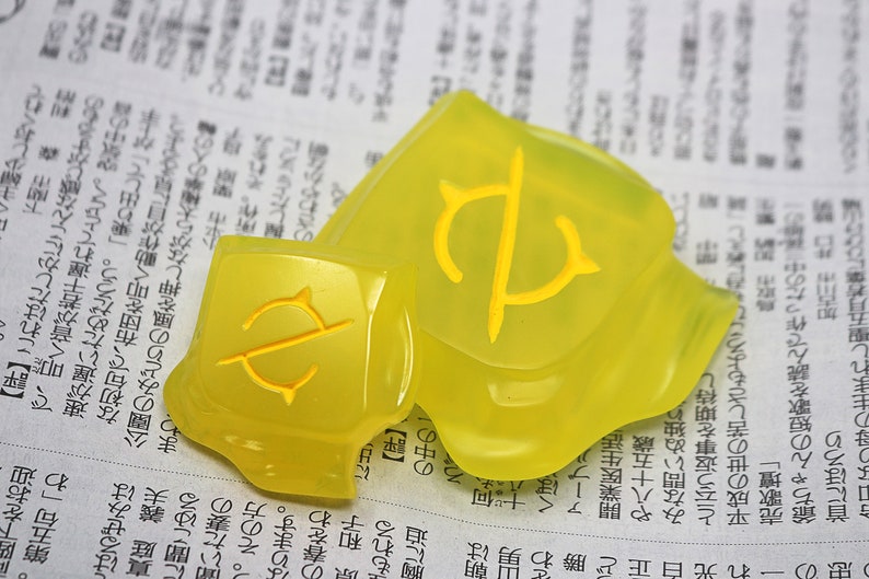 May include: Two translucent yellow decorative pieces with a yellow symbol. The pieces are on a newspaper background. The symbol appears to be a stylized letter or emblem.