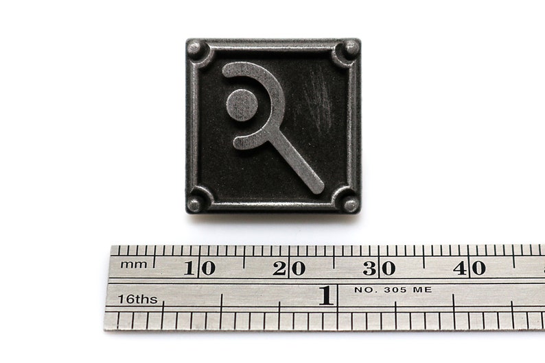 May include: A square, dark gray metal button with a stylized magnifying glass design. The button has rounded corners and small rivets. A ruler with both inches and millimeters is below the button.