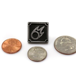 Puede incluir: Un pin de plata con un fondo cuadrado negro y un s&iacute;mbolo de llama plateado. El pin est&aacute; rodeado de tres monedas estadounidenses: un centavo, una moneda de diez centavos y un cuarto de d&oacute;lar.