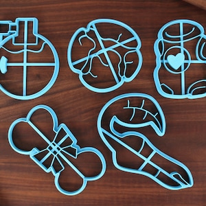 May include: Five blue plastic cookie cutters in different shapes: a circle with a cross inside, a circle with a leaf design, a robot with a heart, a bone with a bow, and a leaf with a stem.