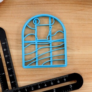 Op de afbeelding: Een lichtblauwe plastic koekjesvormer in de vorm van een raam met een golvend ontwerp. Het raam heeft een afgeronde bovenkant en een rechthoekig frame.