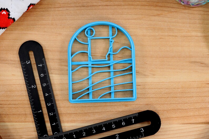 Op de afbeelding: Een lichtblauwe plastic koekjesvormer in de vorm van een raam met een golvend ontwerp. Het raam heeft een afgeronde bovenkant en een rechthoekig frame.