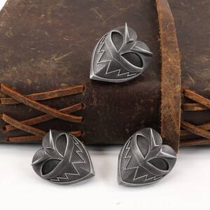 May include: Three silver heart-shaped charms with a stylized face design. The charms are on a brown leather surface.