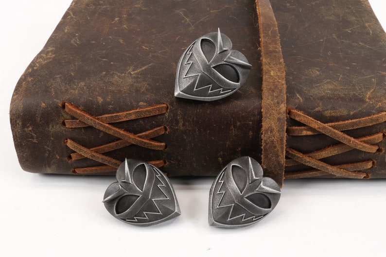 May include: Three silver heart-shaped charms with a stylized face design. The charms are on a brown leather surface.