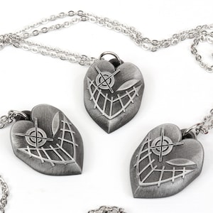 May include: Three silver heart-shaped pendants with a stitched design and a crosshair symbol inside each heart. The pendants are attached to silver chains.