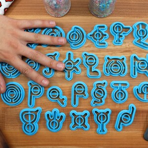 May include: A set of 24 blue plastic cookie cutters shaped like different Pokemon symbols. The cutters are arranged in four rows of six on a wooden surface.