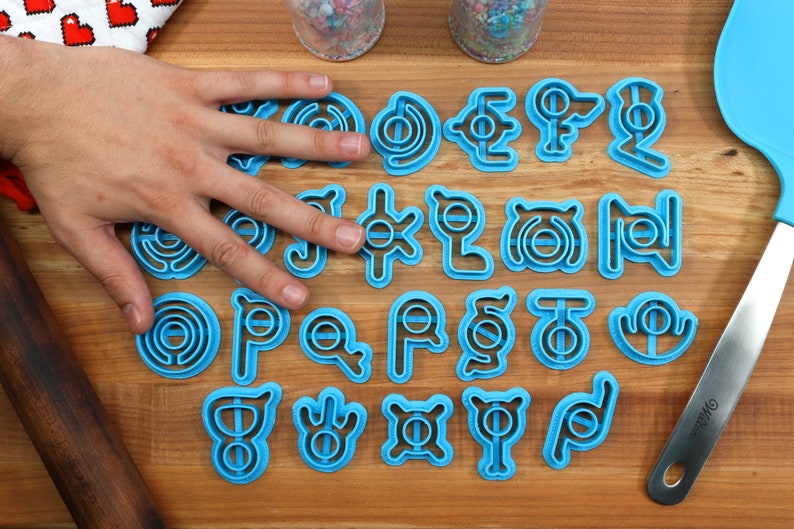 May include: A set of 24 blue plastic cookie cutters shaped like different Pokemon symbols. The cutters are arranged in four rows of six on a wooden surface.