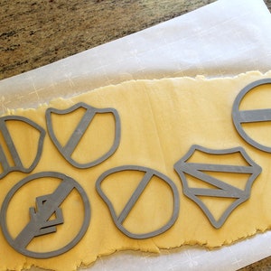 May include: A sheet of cookie dough with various cookie cutters pressed into it. The cutters are in the shapes of shields, circles, and other geometric designs. The dough is a light yellow color and the cutters are a silver color.
