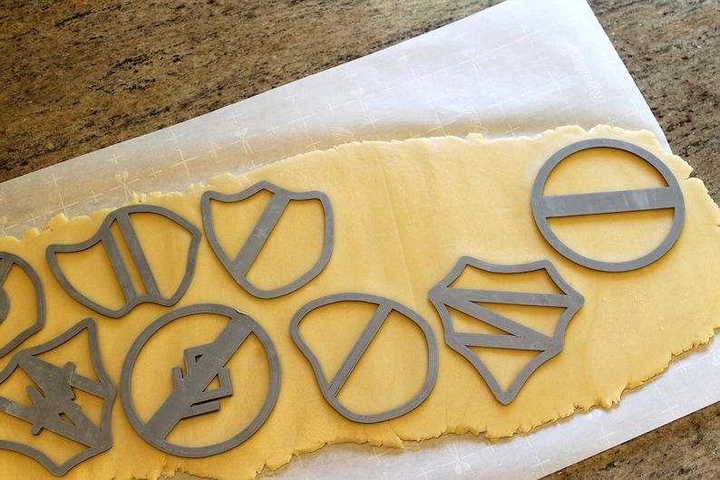 May include: A sheet of cookie dough with various cookie cutters pressed into it. The cutters are in the shapes of shields, circles, and other geometric designs. The dough is a light yellow color and the cutters are a silver color.