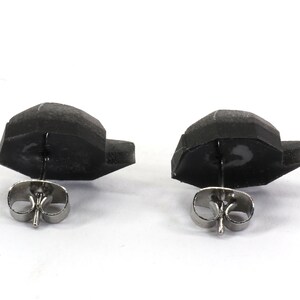 May include: A pair of black stud earrings with silver posts. The earrings are shaped like a small, flat, geometric shape.