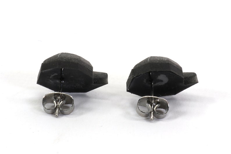 May include: A pair of black stud earrings with silver posts. The earrings are shaped like a small, flat, geometric shape.