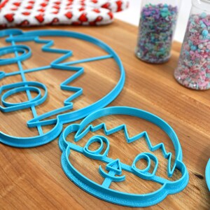 May include: Two blue plastic cookie cutters shaped like a person's face. The larger cutter has a more detailed design with a wavy hairline, eyes, nose, and mouth. The smaller cutter has a simpler design with a wavy hairline, eyes, and a smile.