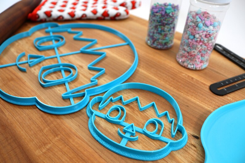 May include: Two blue plastic cookie cutters shaped like a person's face. The larger cutter has a more detailed design with a wavy hairline, eyes, nose, and mouth. The smaller cutter has a simpler design with a wavy hairline, eyes, and a smile.