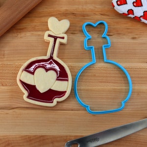 May include: A blue cookie cutter shaped like a bottle with a heart on top, next to a decorated cookie with red and white icing.