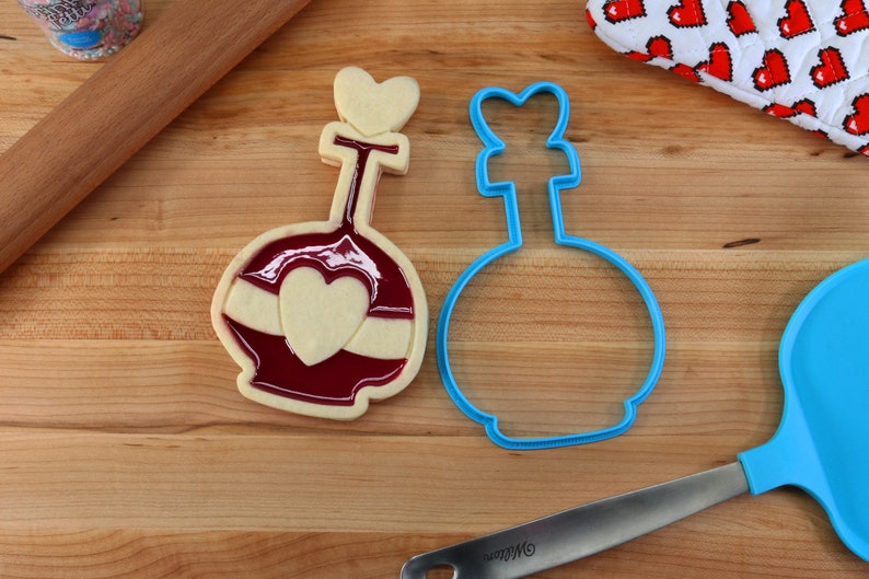 May include: A blue cookie cutter shaped like a bottle with a heart on top, next to a decorated cookie with red and white icing.