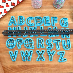 May include: A set of 26 blue plastic alphabet cookie cutters on a wooden surface. The cutters are arranged in alphabetical order. A ruler, rolling pin, and spatula are also visible in the image.