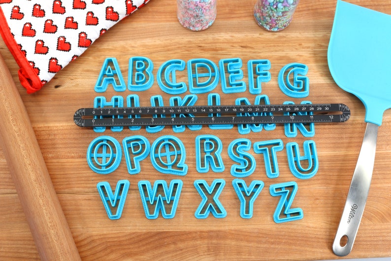 May include: A set of 26 blue plastic alphabet cookie cutters on a wooden surface. The cutters are arranged in alphabetical order. A ruler, rolling pin, and spatula are also visible in the image.