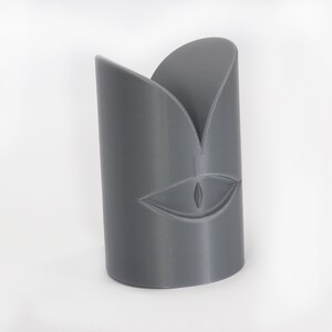 May include: A grey 3D printed sculpture of a face with a single eye and a stylised mouth.