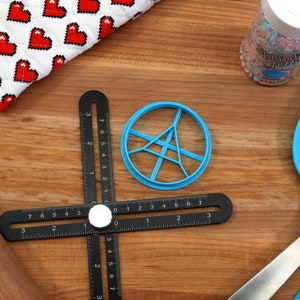 May include: A blue plastic cookie cutter shaped like a circle with a starburst design inside. The cutter is on a wooden surface with a black ruler, a rolling pin, and a blue spatula.