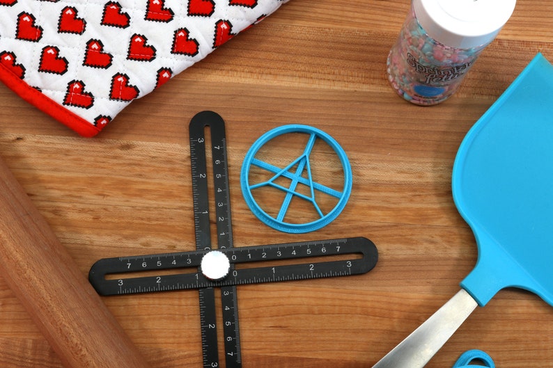May include: A blue plastic cookie cutter shaped like a circle with a starburst design inside. The cutter is on a wooden surface with a black ruler, a rolling pin, and a blue spatula.