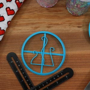 May include: A blue plastic cookie cutter shaped like a stylized letter 'L' with a circular frame. The cutter is on a wooden surface with a ruler and a blue spatula.