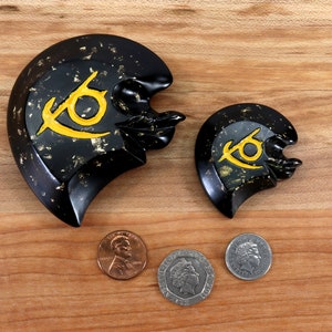 May include: Two black heart-shaped decorative pieces with a gold and yellow eye symbol. The larger piece is on the left and the smaller piece is on the right. Both pieces are on a wooden surface.