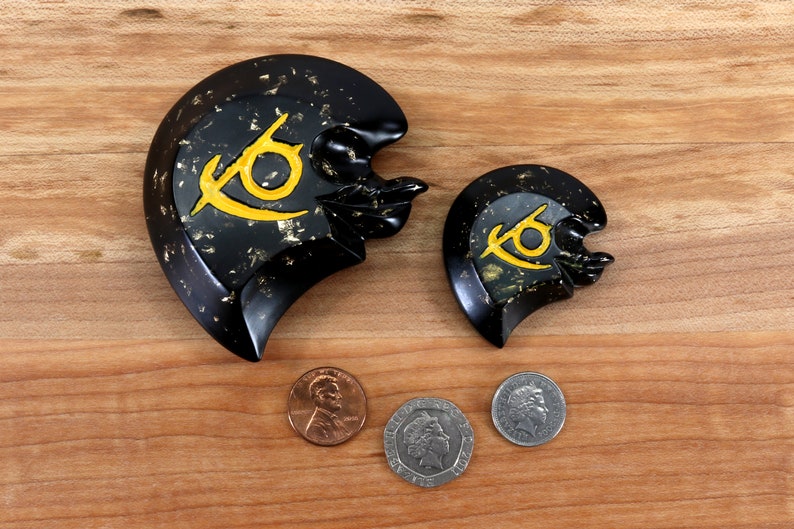 May include: Two black heart-shaped decorative pieces with a gold and yellow eye symbol. The larger piece is on the left and the smaller piece is on the right. Both pieces are on a wooden surface.
