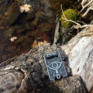 May include: A silver metal pendant featuring a stylised eye design and a triangle pattern. The pendant is resting on a wooden surface with a mossy background.