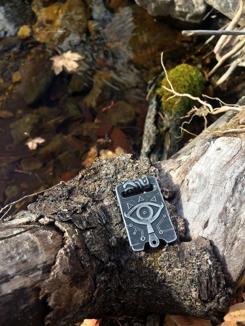 May include: A silver metal pendant featuring a stylised eye design and a triangle pattern. The pendant is resting on a wooden surface with a mossy background.