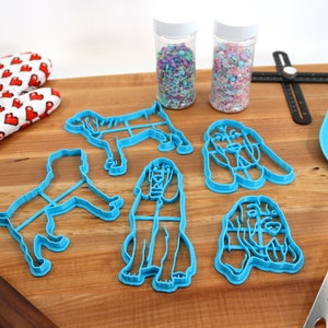 May include: Five blue plastic cookie cutters shaped like bloodhounds, two jars of colorful sprinkles, a rolling pin, a spatula, and a ruler on a wooden surface.