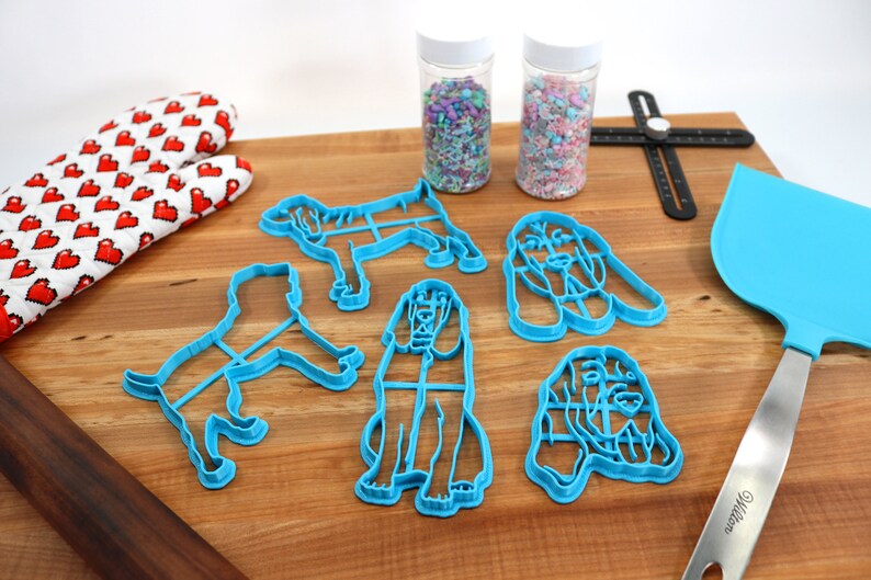 May include: Five blue plastic cookie cutters shaped like bloodhounds, two jars of colorful sprinkles, a rolling pin, a spatula, and a ruler on a wooden surface.