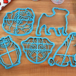 May include: Set of five blue plastic cookie cutters shaped like pugs. The cutters are on a wooden surface.