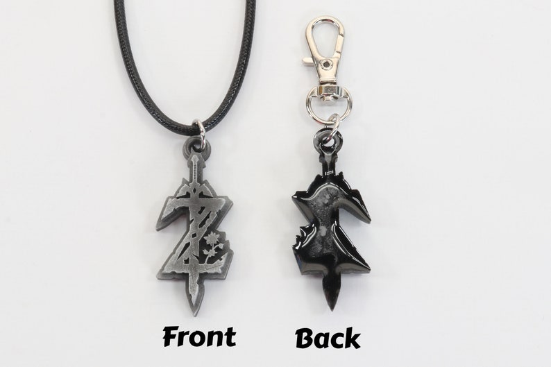 May include: A silver-colored pendant necklace with a black cord and a silver-colored clasp. The pendant is a stylized sword with a design on the front and a black finish on the back. The pendant is also available as a keychain with a silver-colored clip.