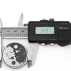 May include: A silver-colored metal pendant with a yin-yang design featuring two stylized dragons. The pendant is being measured with a digital caliper, which reads 34.97 mm.