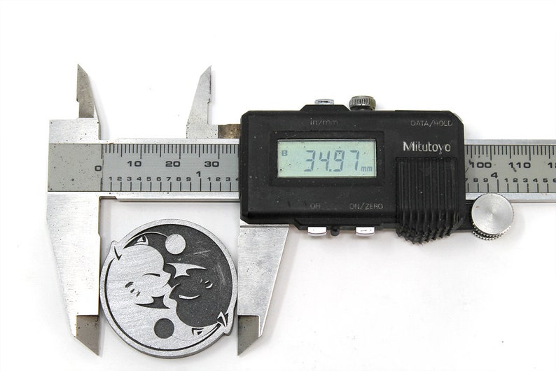 May include: A silver-colored metal pendant with a yin-yang design featuring two stylized dragons. The pendant is being measured with a digital caliper, which reads 34.97 mm.