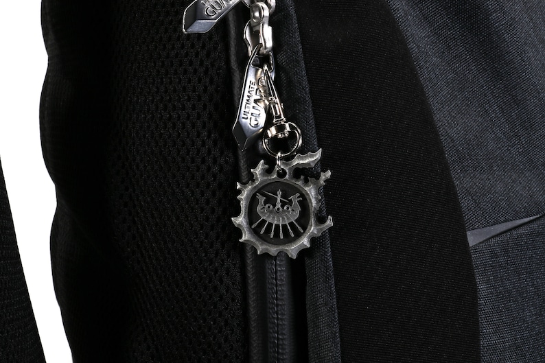 Op de afbeelding: Een zwarte rugzak met een zilveren rits met een metalen hanger met een schipontwerp. De hanger is bevestigd aan een zilveren clip.