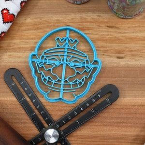 May include: Blue plastic cookie cutter shaped like a cartoon character with a crown, mustache, and a mischievous grin.