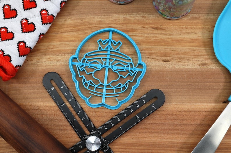 May include: Blue plastic cookie cutter shaped like a cartoon character with a crown, mustache, and a mischievous grin.