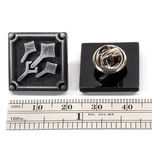 May include: A silver-colored metal pin with a square design featuring a stylized image of three arrows pointing in different directions. The pin is attached to a black backing card and is shown next to a ruler for size reference.