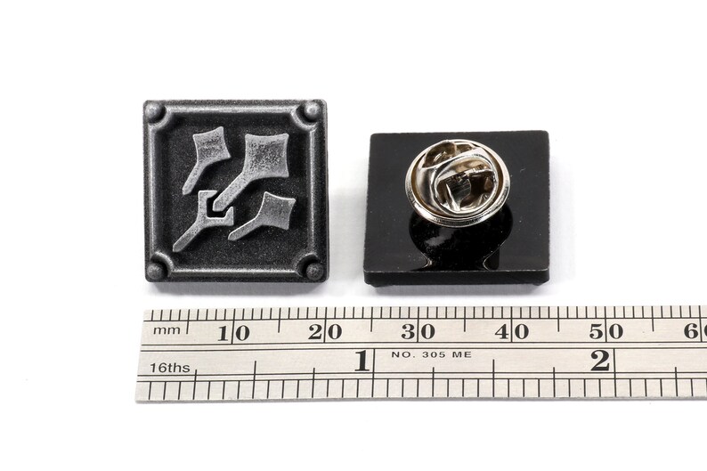 May include: A silver-colored metal pin with a square design featuring a stylized image of three arrows pointing in different directions. The pin is attached to a black backing card and is shown next to a ruler for size reference.