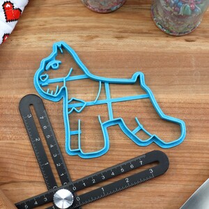 May include: Blue plastic cookie cutter shaped like a miniature schnauzer dog. The cutter is on a wooden surface with a ruler and other baking tools.