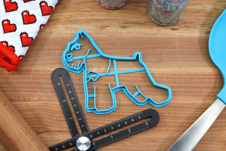 May include: Blue plastic cookie cutter shaped like a miniature schnauzer dog. The cutter is on a wooden surface with a ruler and other baking tools.