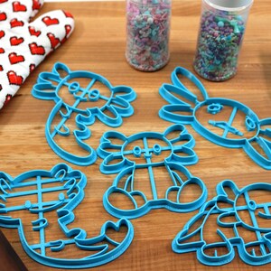 May include: A set of five blue plastic cookie cutters shaped like axolotls. The cutters are on a wooden surface with a red and white oven mitt, a rolling pin, and two jars of sprinkles.