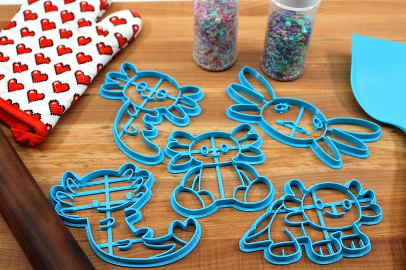 May include: A set of five blue plastic cookie cutters shaped like axolotls. The cutters are on a wooden surface with a red and white oven mitt, a rolling pin, and two jars of sprinkles.