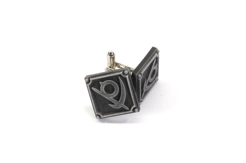May include: A pair of silver cufflinks with a diamond-shaped design featuring a stylized symbol of a sword and a circle.