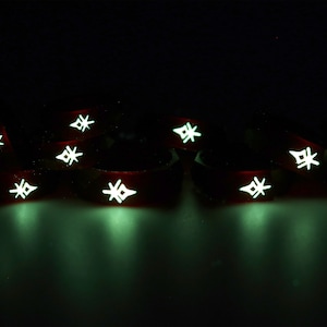 May include: A group of eight red glow-in-the-dark silicone bracelets with a white starburst design. The bracelets are arranged in a semi-circle on a dark surface.