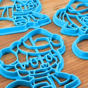 May include: Three blue plastic cookie cutters shaped like cartoon characters. The cutters are on a wooden surface.